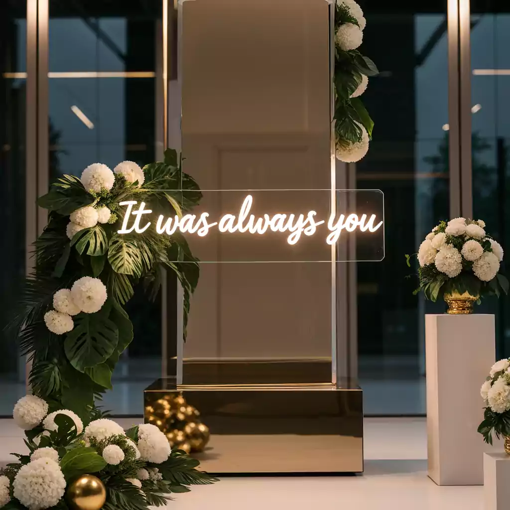 Luxurious wedding decor featuring a gold pedestal, lush white floral arrangements, and a glowing neon sign that reads It was always you.