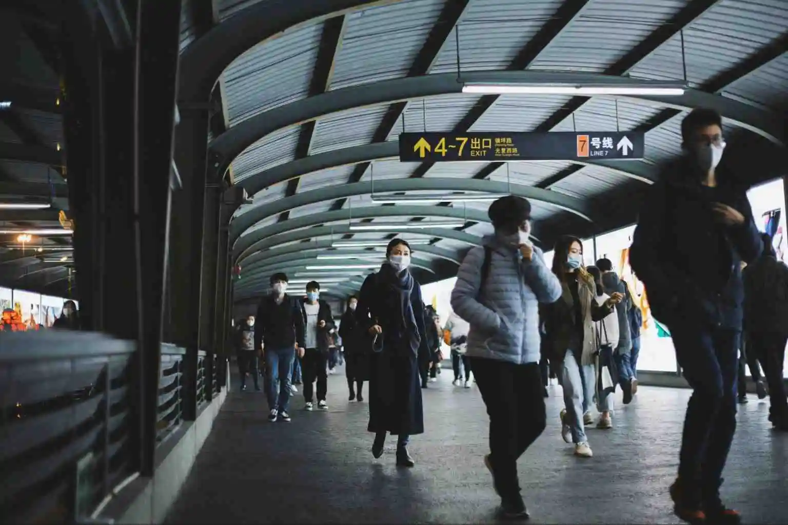 People wearing face masks walking along a spacious covered walkway with directional signage and curved roof, symbolising logistical coordination at large events.